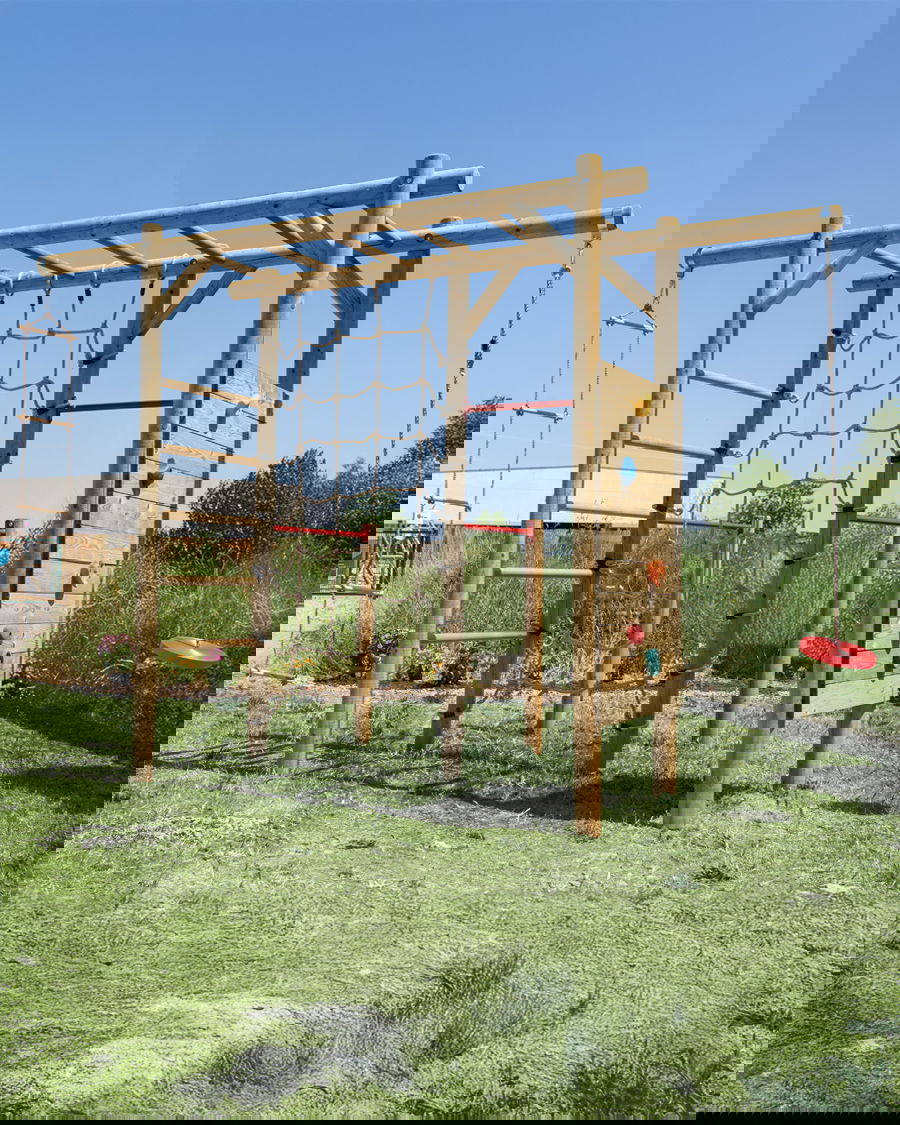 Holzspielplatz Kletterstruktur mit Seilnetz, Kletterwand und Schwung auf dem grasbewachsenen Feld unter klarem blauem Himmel. Perfekt für Kinderaktivitäten im Freien.
