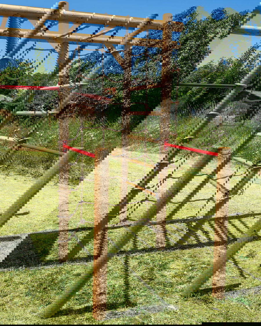 Holzkletternstruktur im Outdoor -Spielplatz mit Seil- und Leiterelementen, die in einem Grasparkbereich unter einem klaren blauen Himmel spielt.